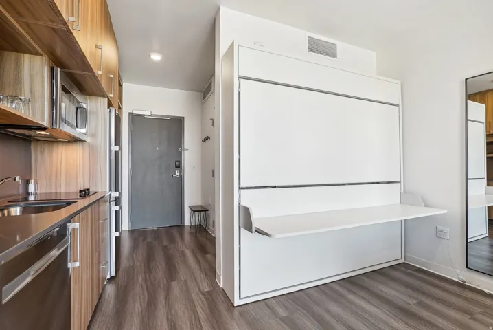 Compact living area featuring a built-in wall bed with an integrated desk, wood-finish flooring, and a stainless steel kitchen with a microwave and refrigerator
