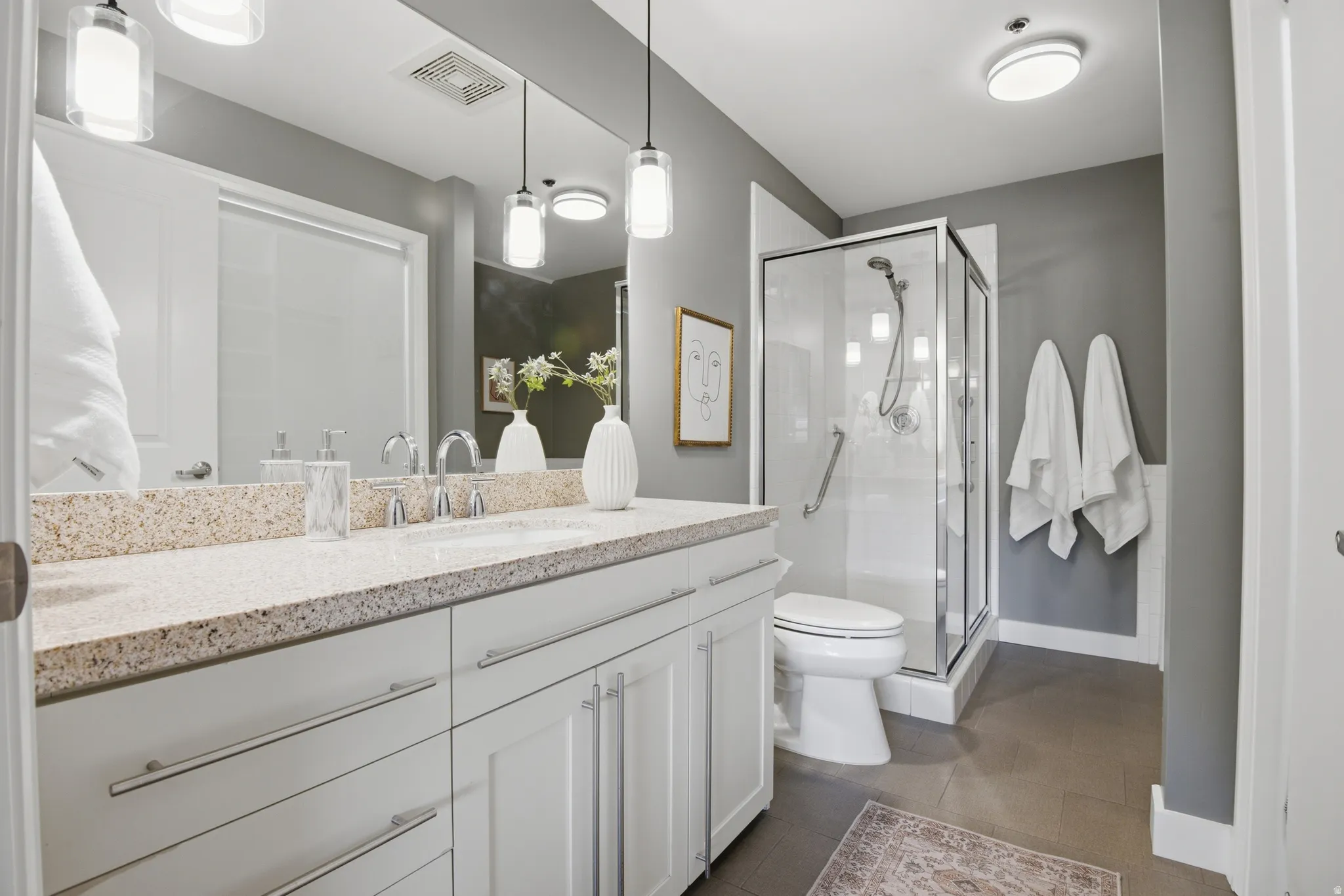 Bathroom featuring a dual vanity with a speckled countertop, undermount sinks, white cabinetry with chrome hardware, and a glass-enclosed shower with a grab bar
