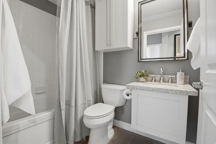 Bathroom featuring a white subway tile bath/shower surround, a white vanity with a speckled countertop, a rectangular framed mirror, and a white wall-mounted cabinet