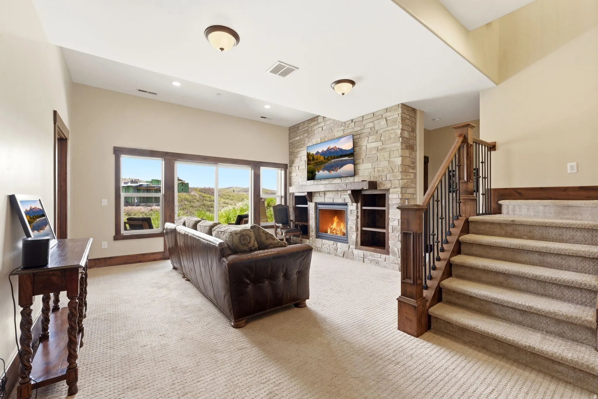 Spacious living area featuring a stone fireplace with integrated shelving, large windows, recessed lighting, and a carpeted staircase with wood and iron railings