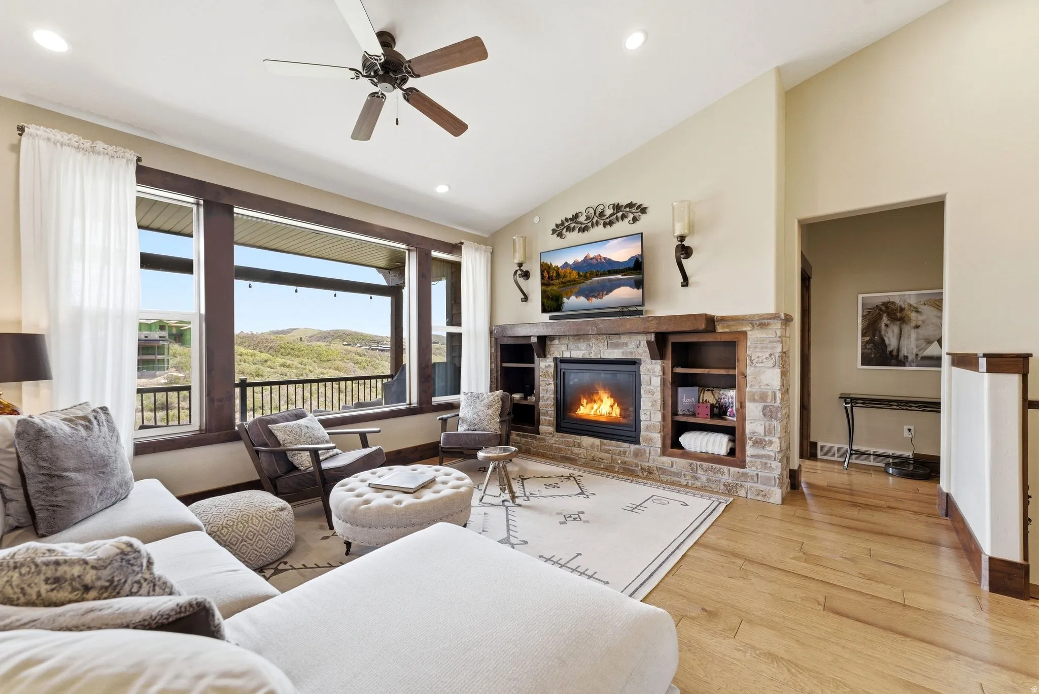 Spacious living area featuring expansive windows, a stone-clad fireplace with a wood mantel, built-in shelving, vaulted ceilings, and wood-finish flooring