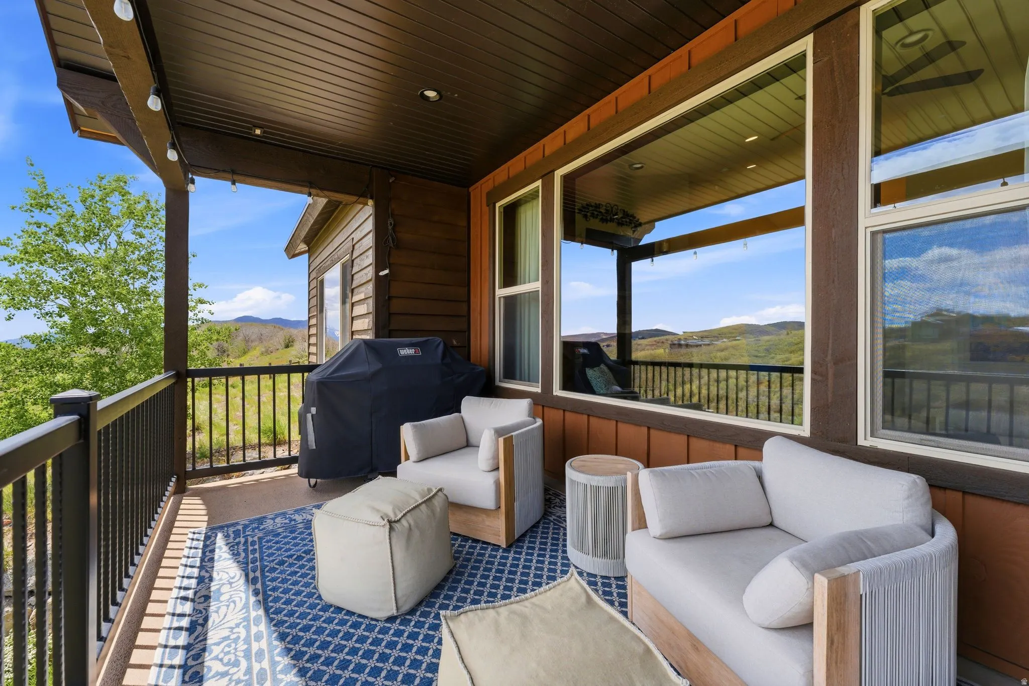 Covered outdoor patio featuring extensive mountain scenery, a dark wood-finish ceiling, pendant lighting, large windows, and a black metal railing