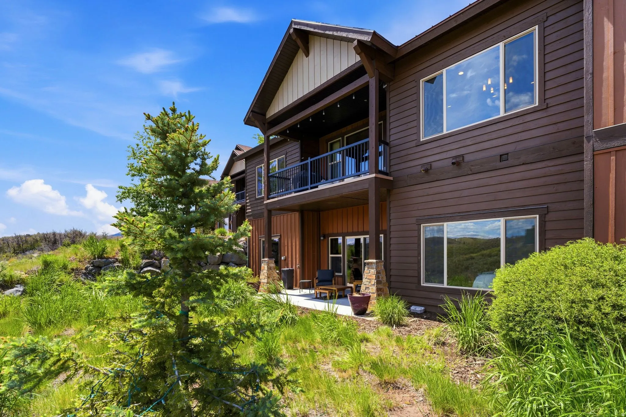 Brown siding facade featuring a top-level balcony with black railings, a lower-level concrete patio, and large windows