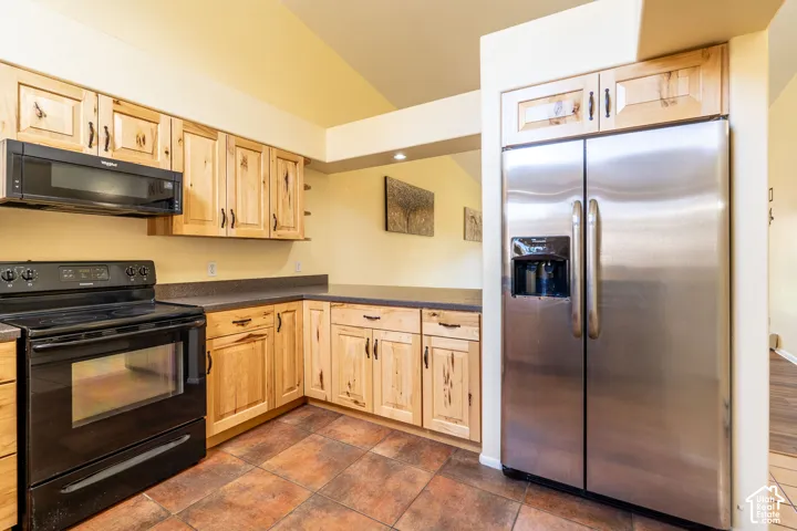 Kitchen featuring natural wood cabinetry, dark countertops, black appliances, a stainless steel refrigerator, and tiled flooring