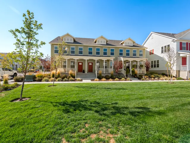 Expansive green lawn with mature trees fronting a two-story residence featuring a covered porch, red entry doors, and symmetrical window placements