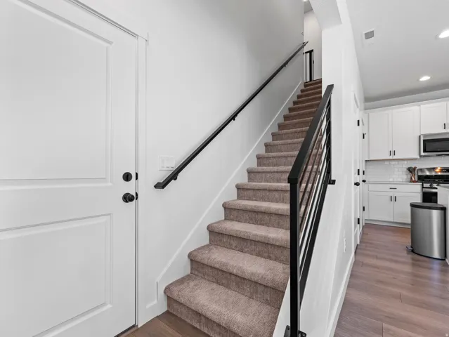 Carpeted staircase with black metal railings, wood-finish flooring, white walls, and a white paneled door