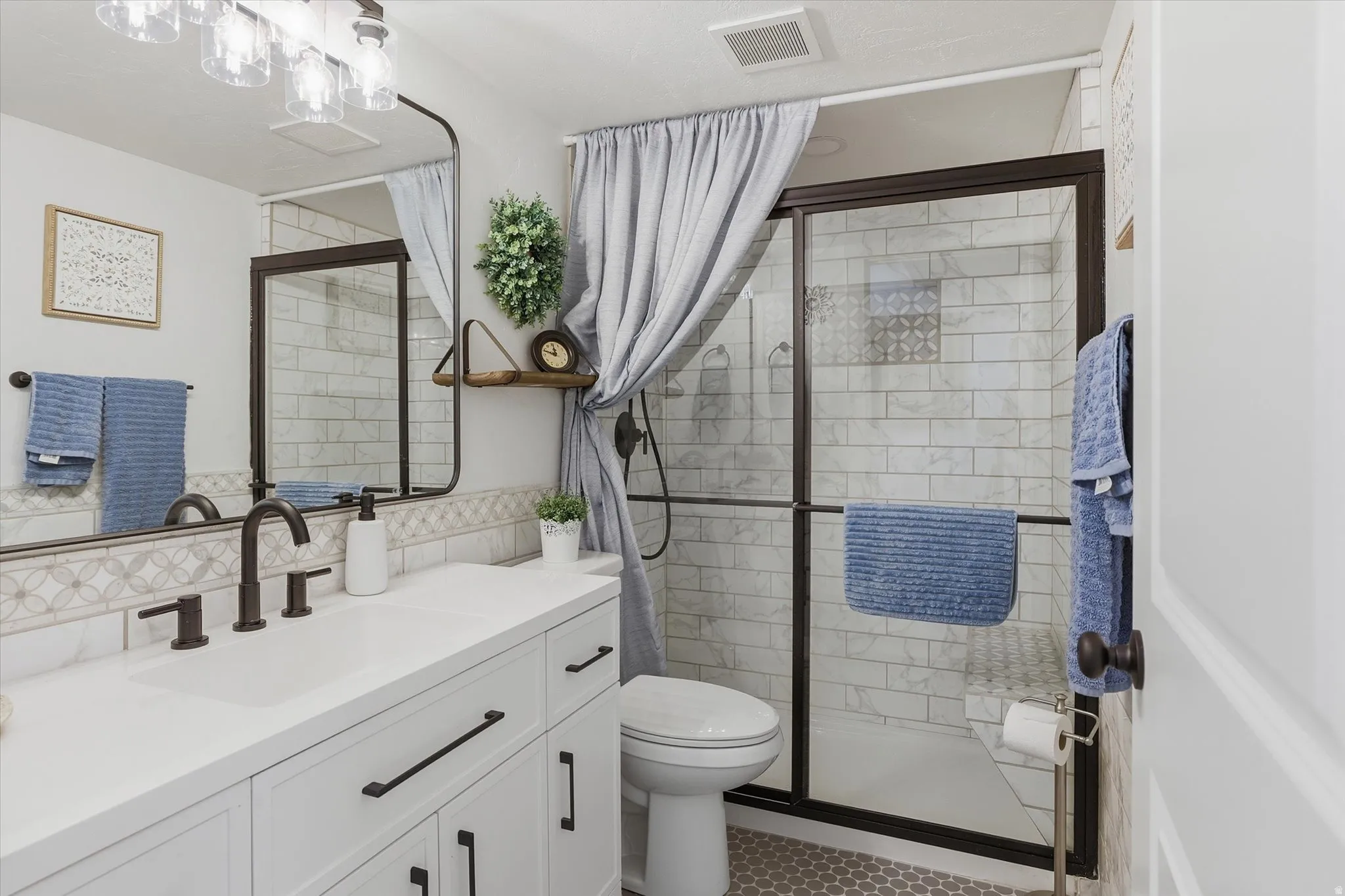 Bathroom featuring a white vanity with a solid surface countertop, bronze fixtures, a framed mirror, and a glass-enclosed shower with subway tile surround and a decorative accent tile