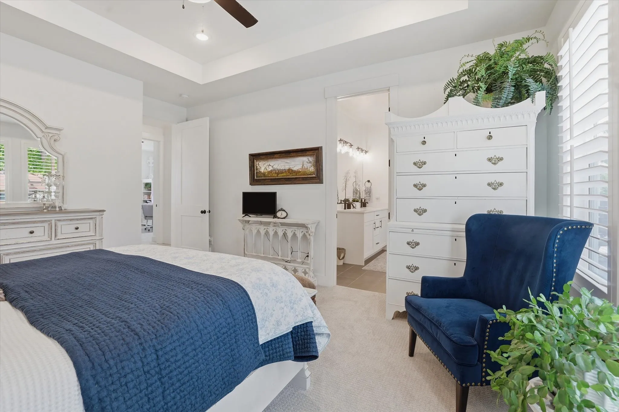 Carpeted room featuring a ceiling fan, tray ceiling, white plantation shutters, and an open doorway revealing a bathroom with a vanity and wall-mounted lighting