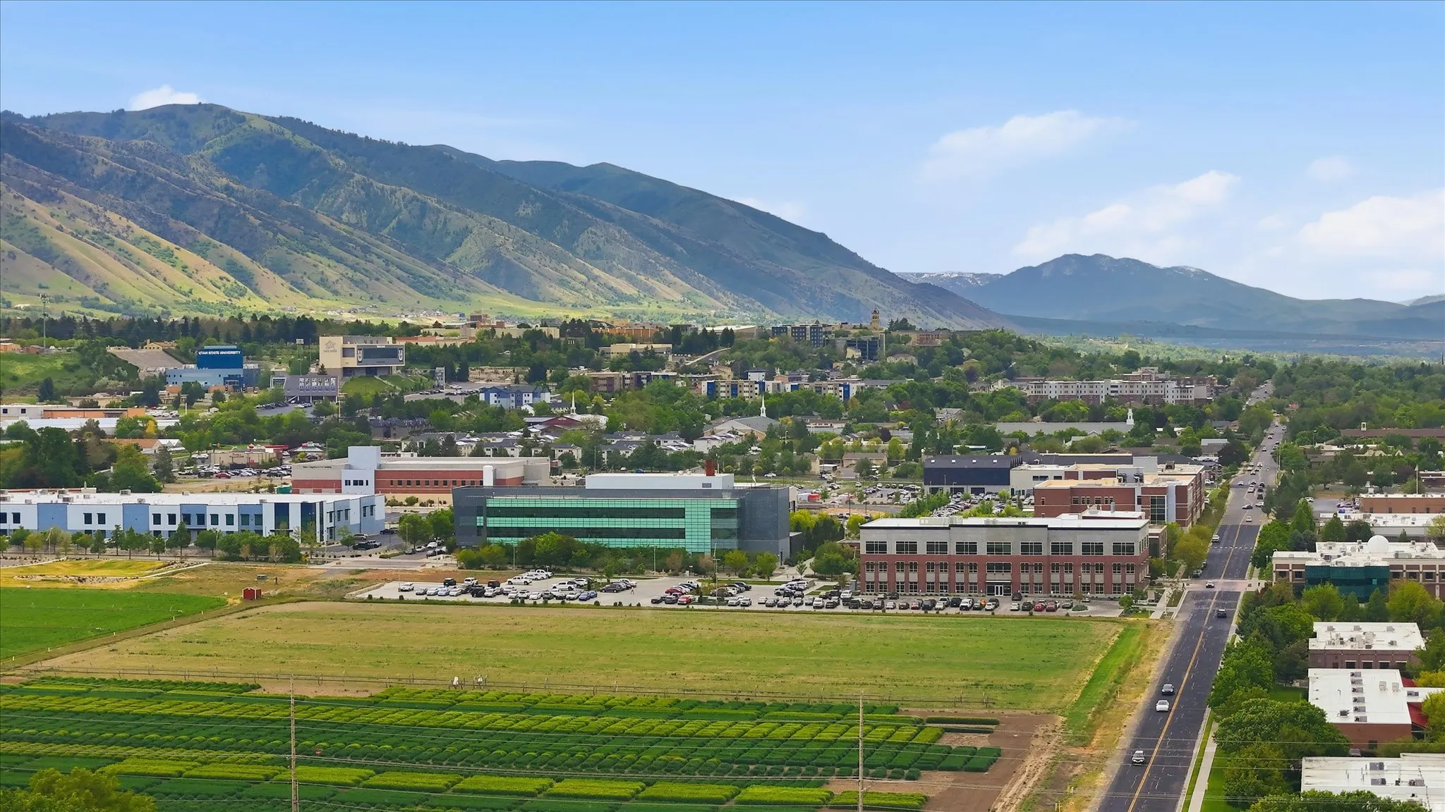 Expansive aerial perspective showcasing a mountainous backdrop, urban development, agricultural fields, and a multi-lane roadway
