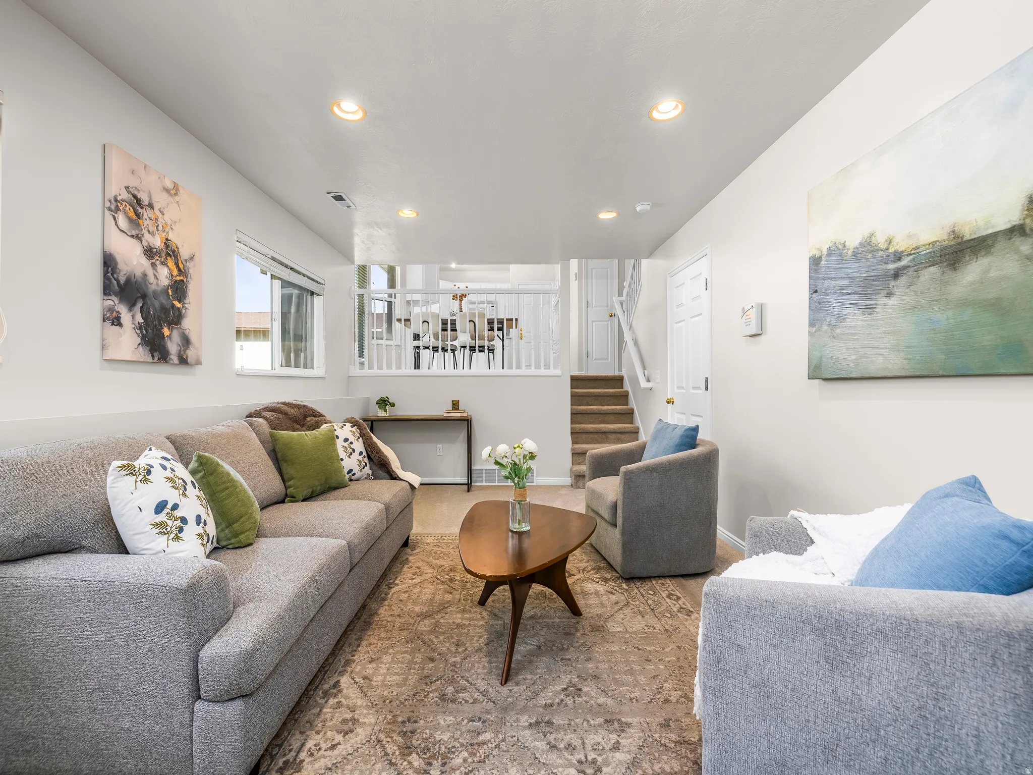 Spacious living area featuring light-toned walls, recessed lighting, wood-finish flooring with an area rug, and an open stairwell with white railings