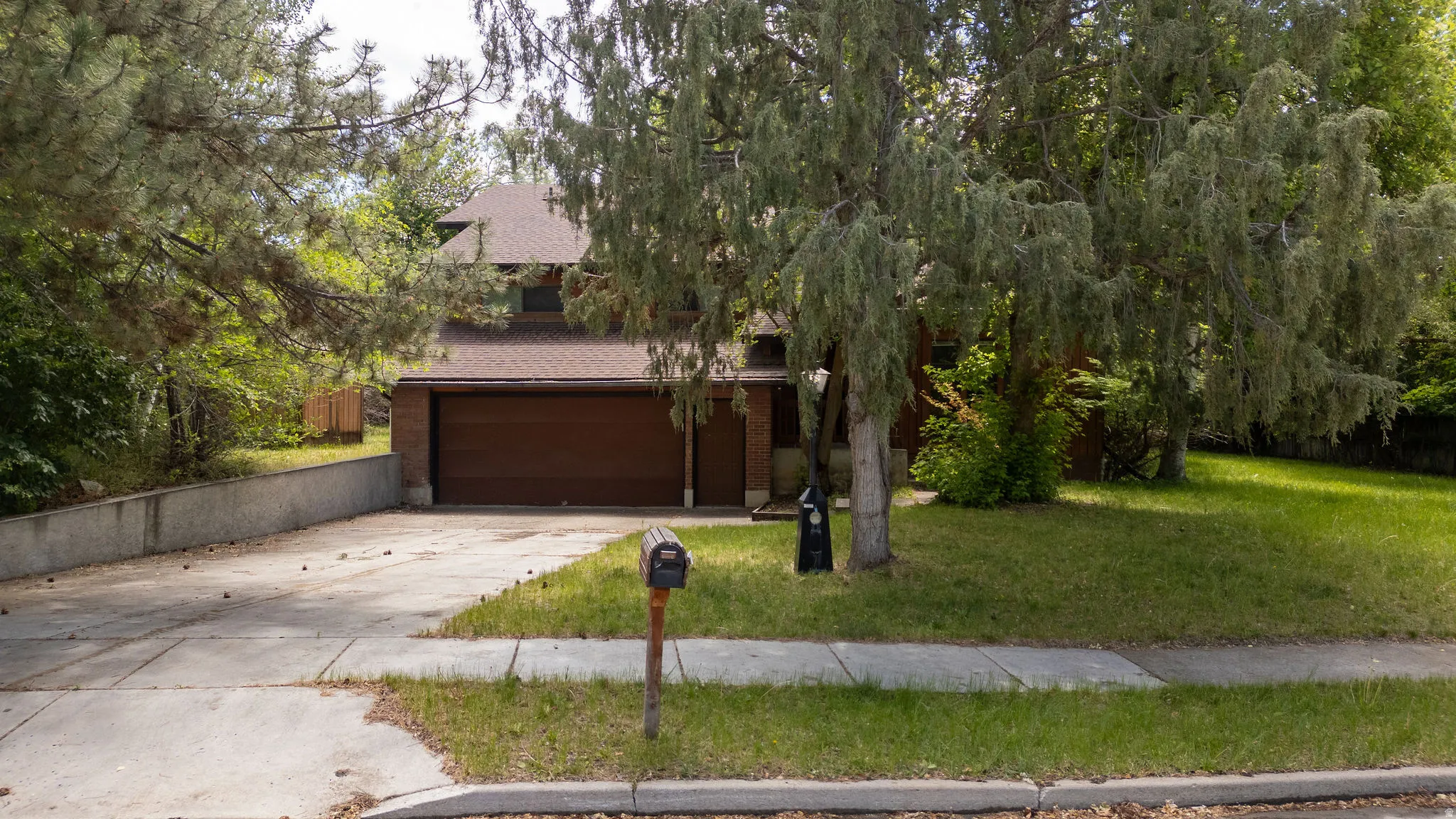 Brick exterior home featuring a concrete driveway, two-car garage with wood-finish door, shingle roof, and mature trees surrounding the property