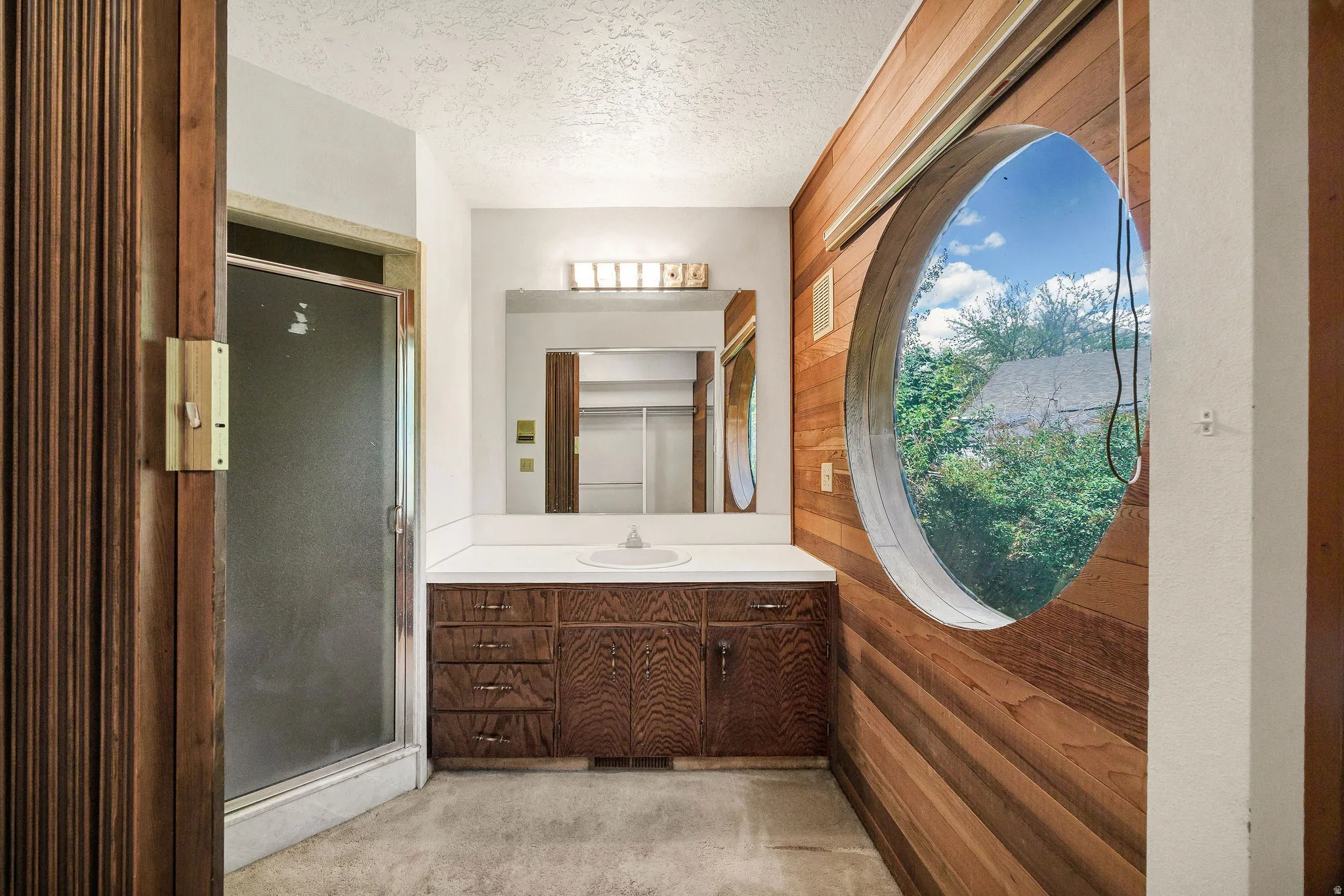 Bathroom featuring a distinctive circular window, wood plank wall paneling, a wood-finish vanity with a white countertop and integrated sink, a large wall-mounted mirror, and a shower enclosure