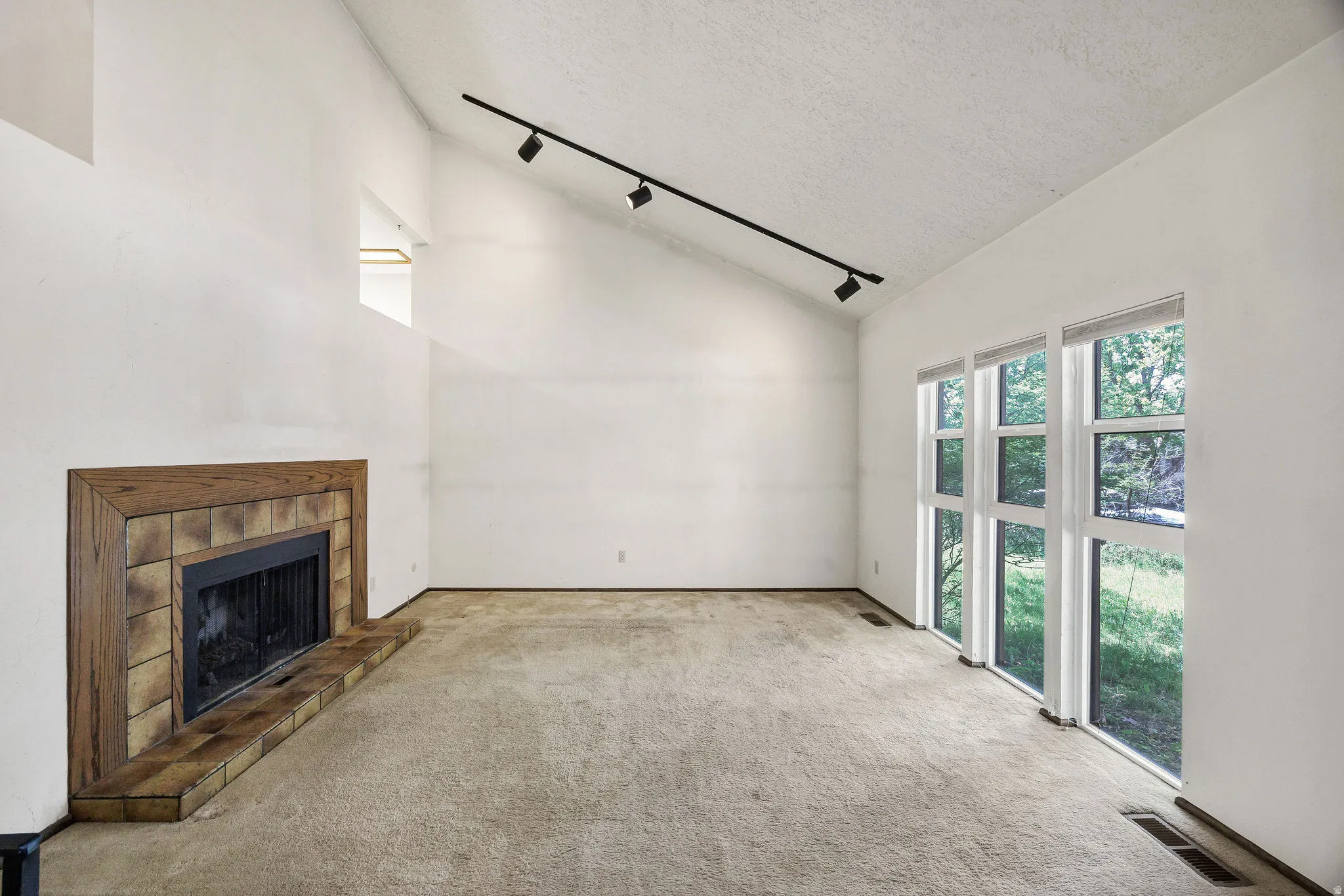 Vaulted ceiling with track lighting, floor-to-ceiling windows, wood-framed fireplace with a tiled surround and hearth, and carpeted flooring