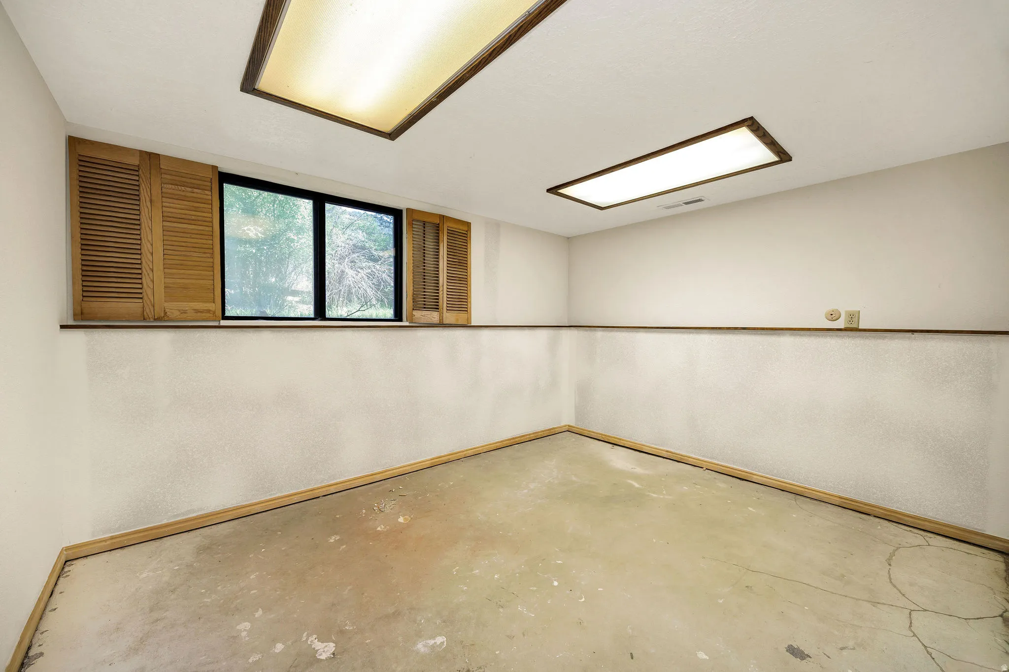Windowed room with a concrete floor, featuring two rectangular ceiling light fixtures and natural wood louvered shutters