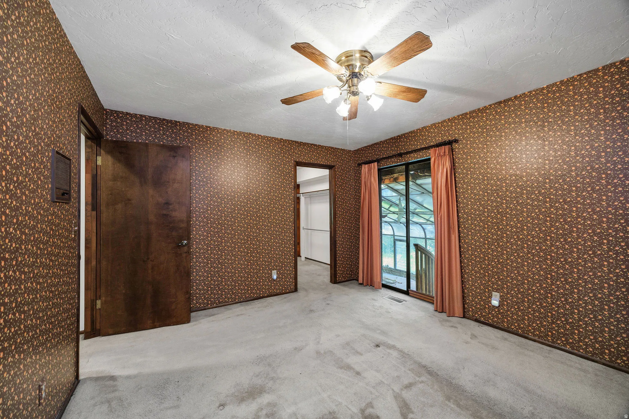 Carpeted room featuring a ceiling fan with light fixture, patterned wallpaper, a wood-finish door, and a sliding glass door with exterior access