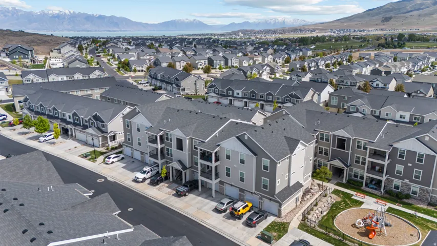Modern multi-family buildings featuring balconies and attached garages, surrounded by a residential neighborhood with distant mountains and a body of water