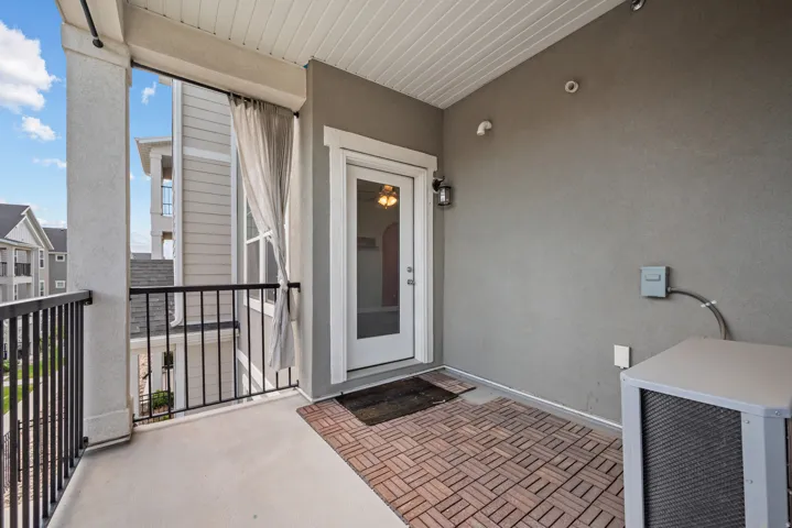 Covered balcony featuring concrete flooring, a partial brick-patterned mat, black metal railings, and a white glass-paneled door