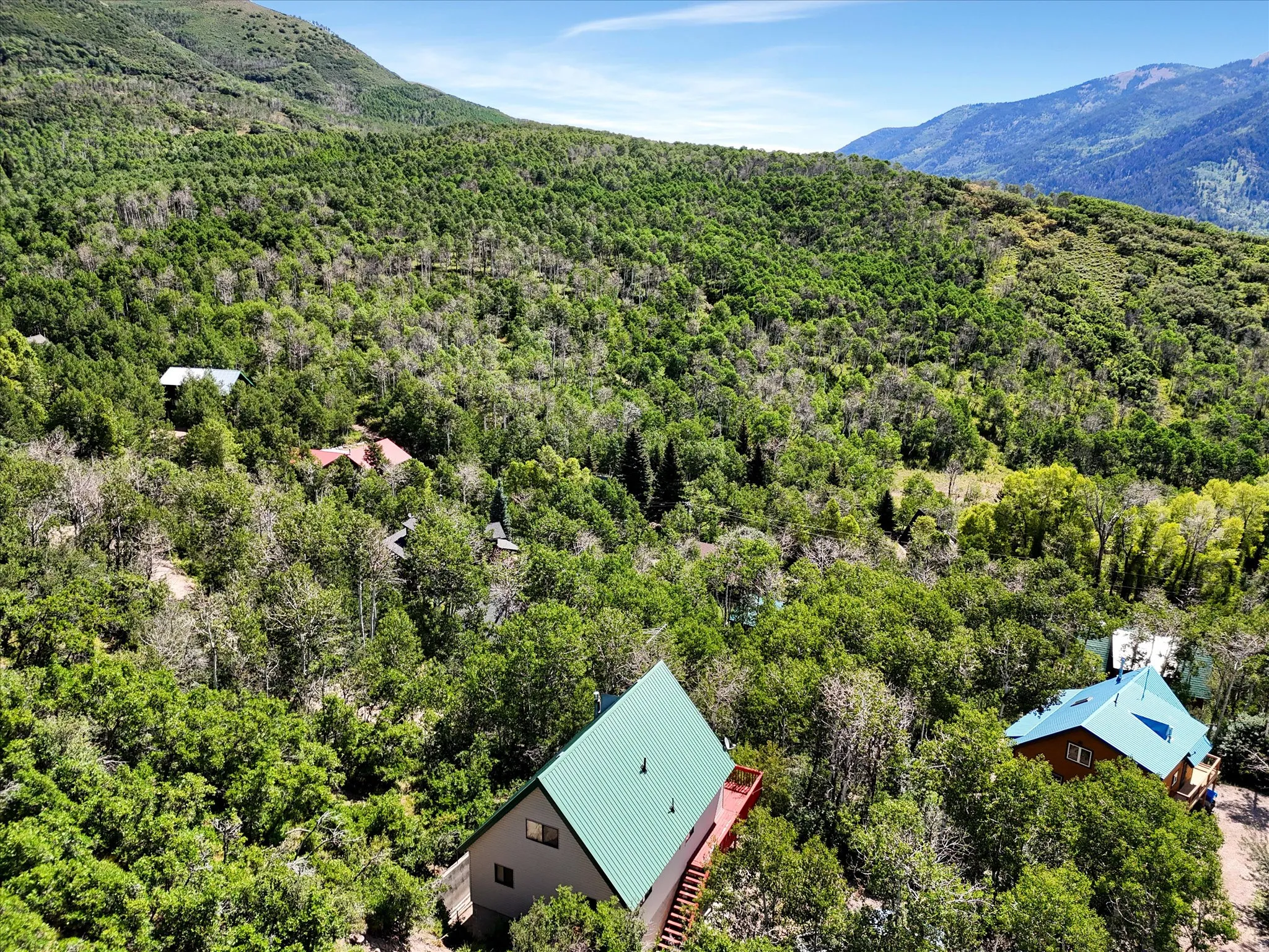 A-frame property featuring a green metal roof, a wood deck, and exterior stairs, surrounded by dense natural landscaping