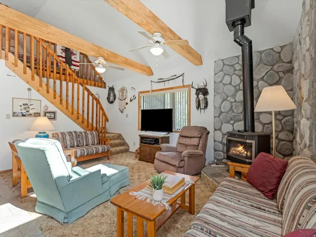 Open living space featuring a natural wood staircase with balusters, exposed ceiling beams, and a rock-faced wall with a wood-burning stove