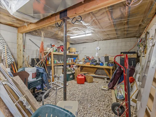 Unfinished basement space featuring exposed wood beams, concrete block walls, a gravel floor, and visible utility runs including ductwork and electrical conduit