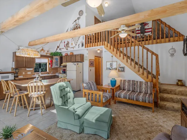 Open-concept living area featuring vaulted ceilings with exposed wood beams, a prominent wooden staircase leading to an upper loft, and a kitchen area with wood-finish cabinetry and a breakfast bar