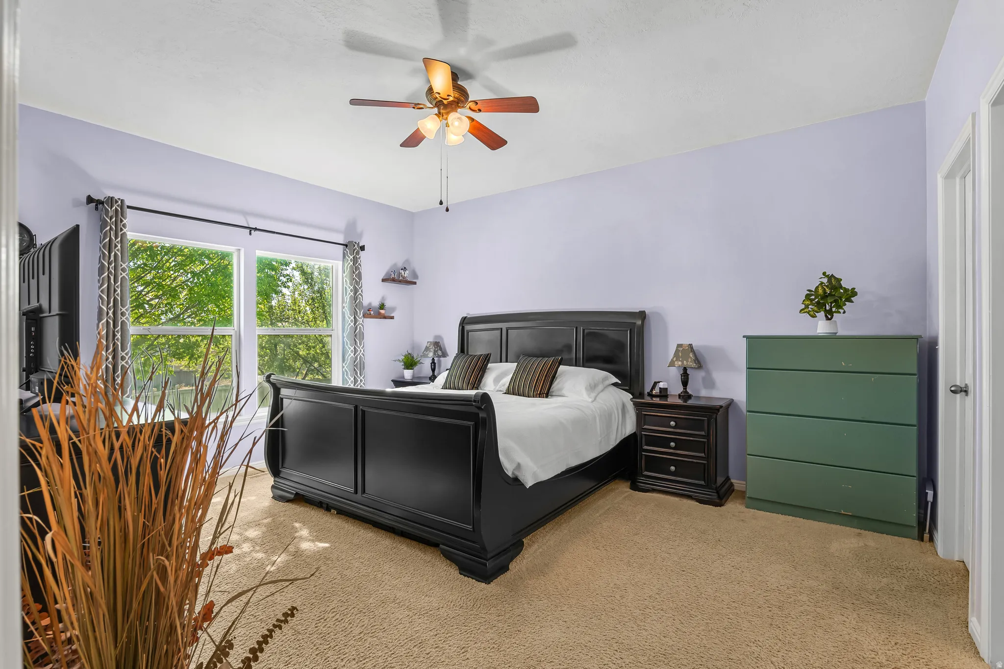 Spacious bedroom featuring carpet flooring, natural light from a large window, a ceiling fan with integrated lighting, and two wall-mounted shelves