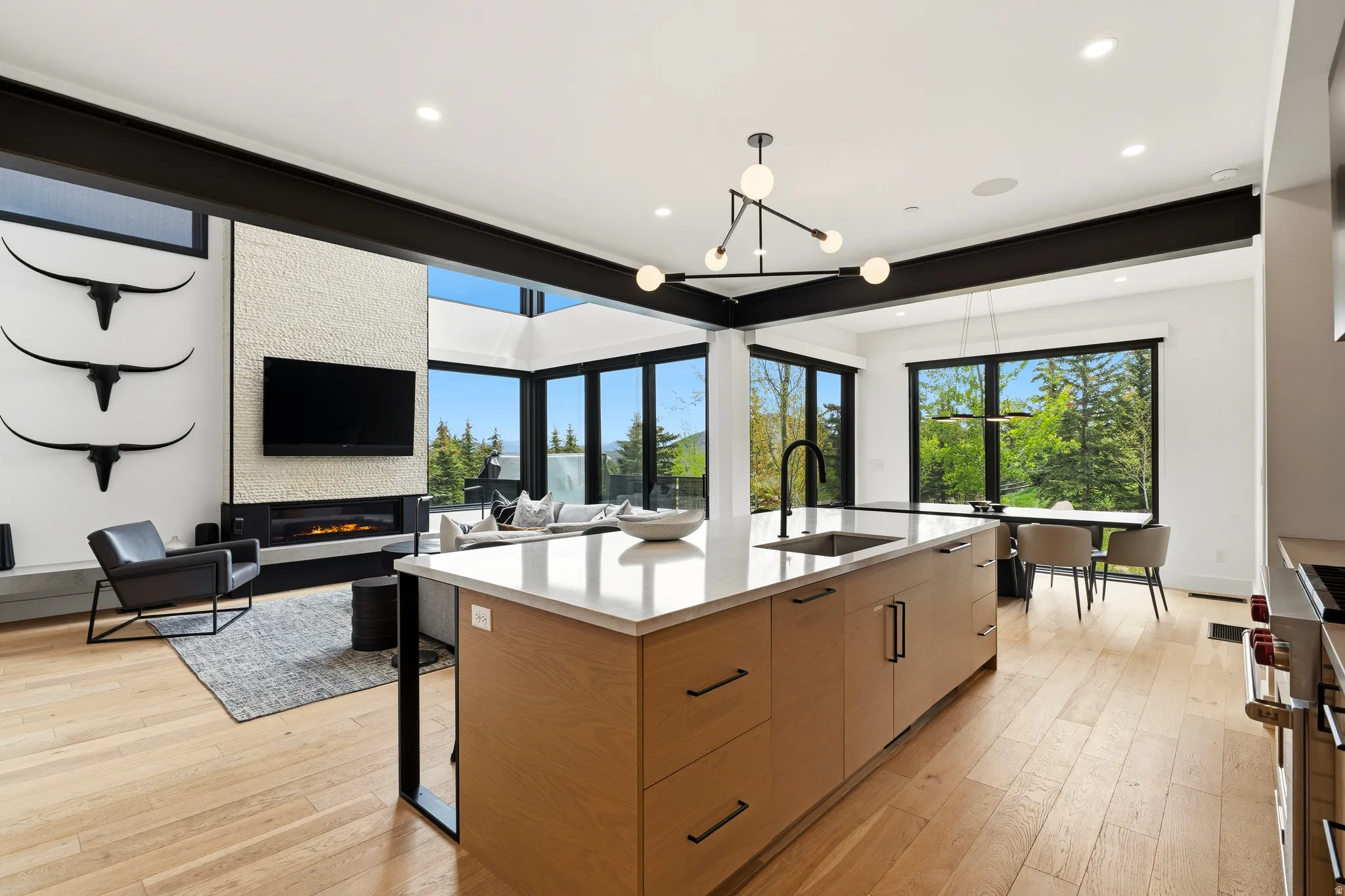 Open-concept living space featuring a central island with a sink, light wood-finish flooring, and large black-framed windows offering exterior views