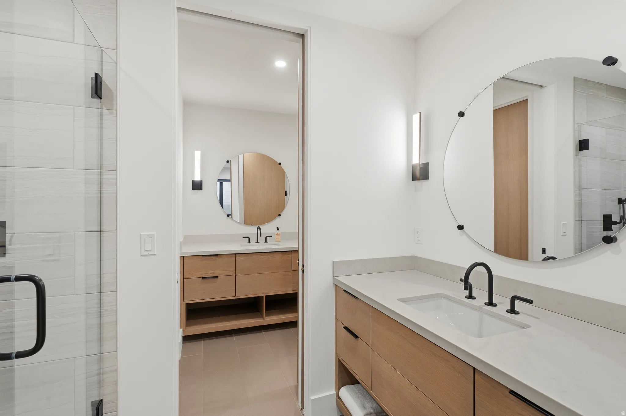 Contemporary bathroom featuring light wood-finish dual vanities with an integrated sink, matte black fixtures, and a large circular mirror
