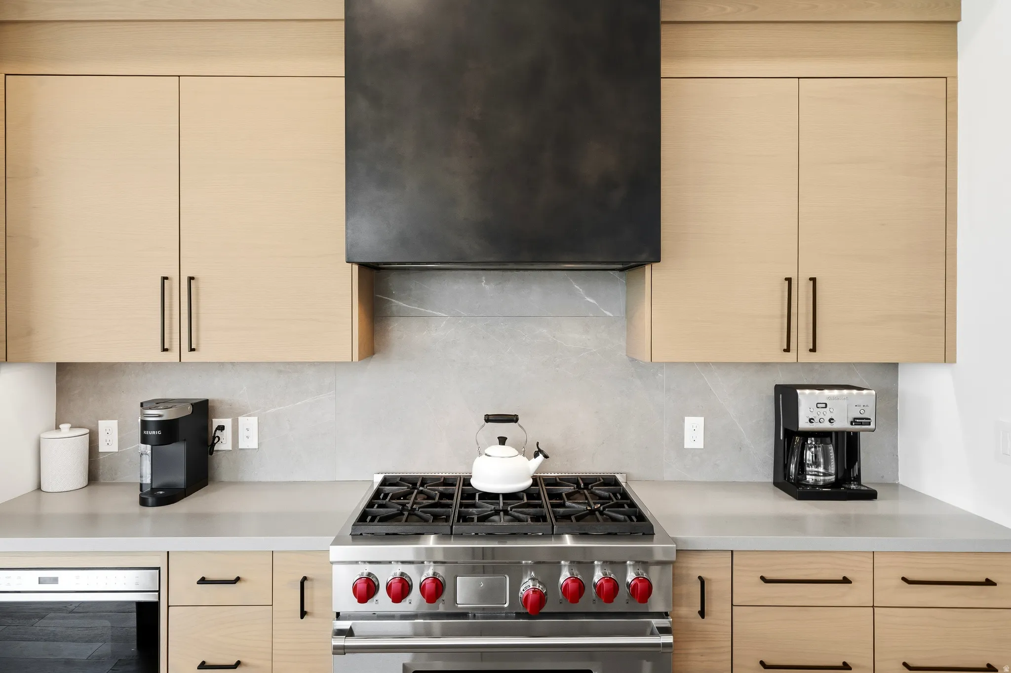 Gourmet kitchen features a professional-grade stainless steel range with red accent knobs, a dark bronze range hood, and light wood-finish cabinetry with black hardware