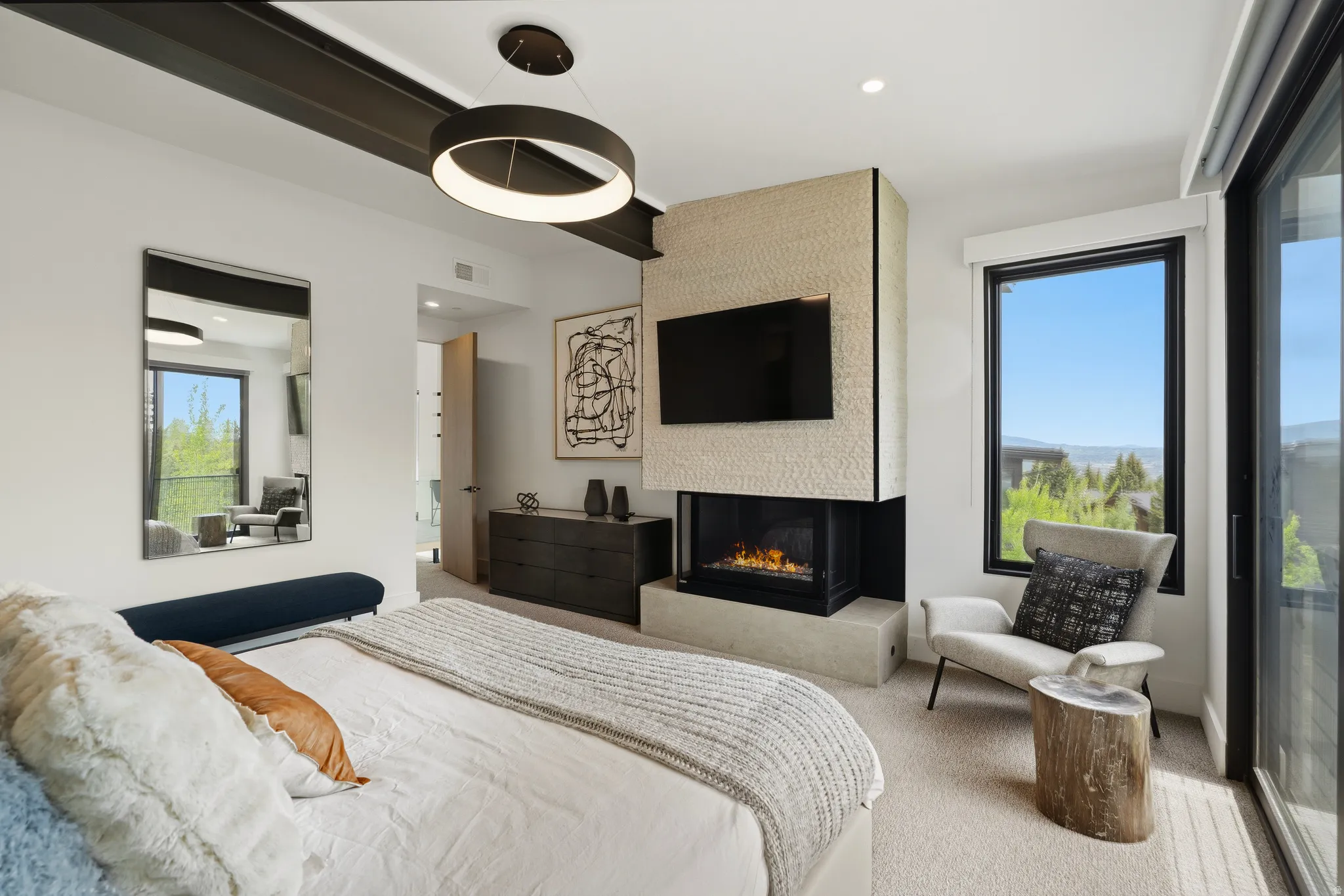 Modern bedroom featuring a stone-clad fireplace, integrated television, and a large window