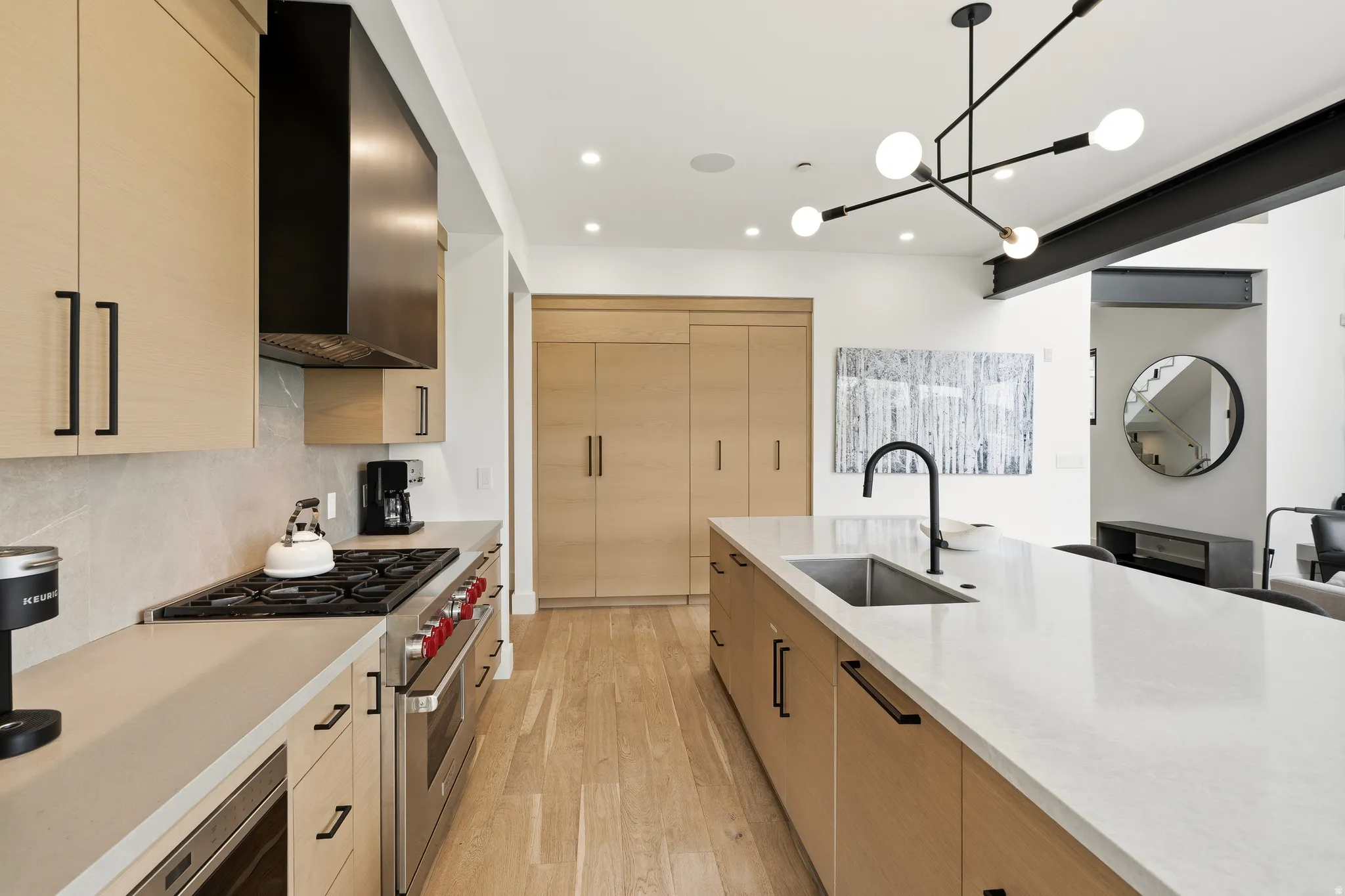 Contemporary kitchen featuring light wood-finish cabinetry, a large center island with an undermount sink and matte black faucet, integrated stainless steel appliances, and wood-finish flooring
