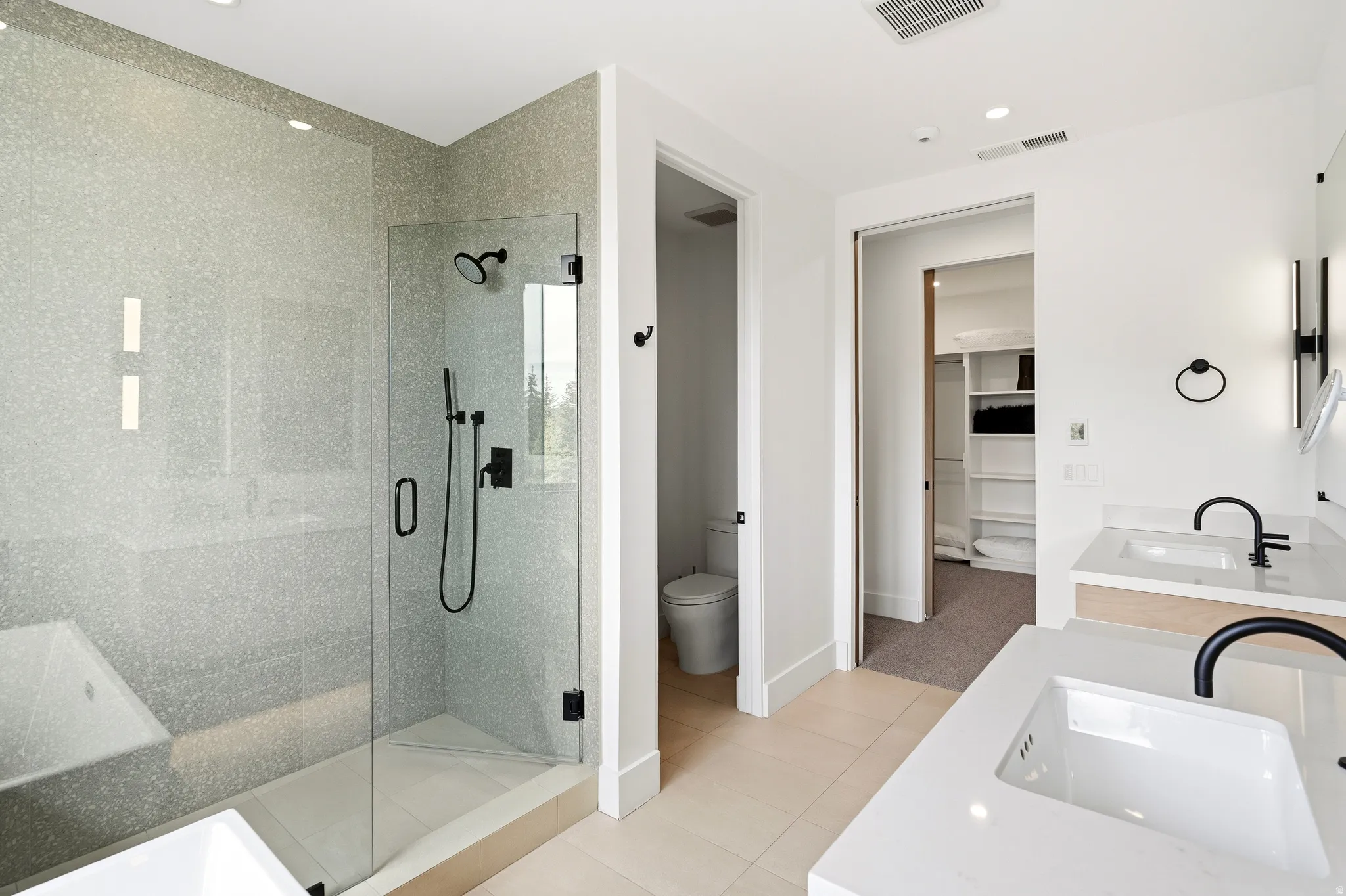 Spacious bathroom featuring a glass-enclosed shower with matte black fixtures, dual vanities with white countertops, and a separate water closet