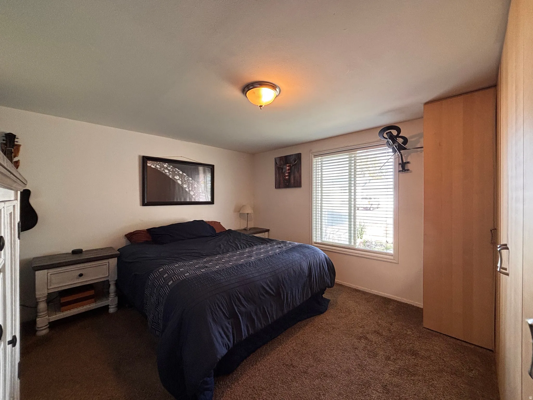 Bedroom featuring light-toned walls, brown carpet flooring, and a large window with white blinds