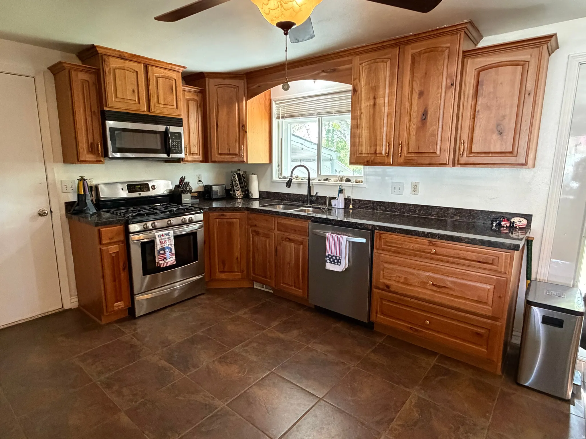 Kitchen featuring wood cabinetry, stainless steel appliances, dark stone-finish countertops, tile flooring, and a ceiling fan with light fixture