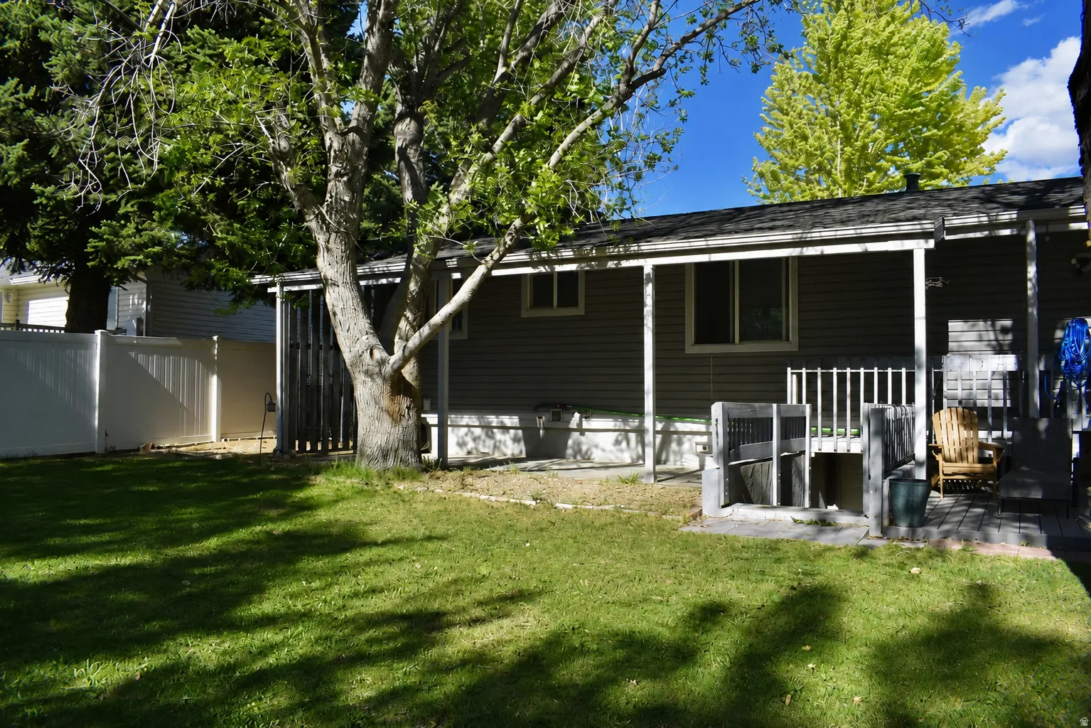 Expansive rear yard featuring a covered patio with railing, mature trees, manicured lawn, and a white privacy fence