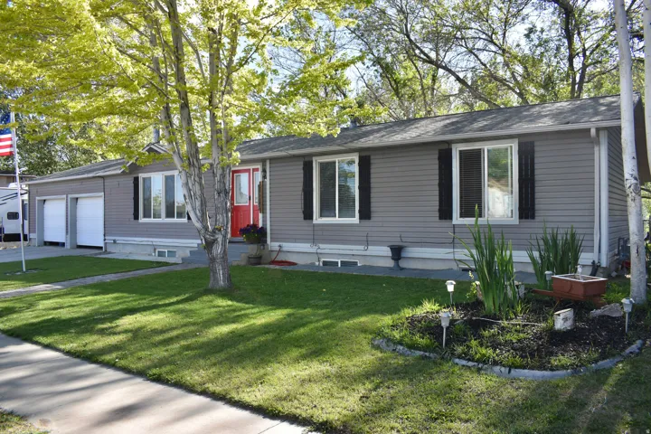Ranch-style exterior with gray horizontal siding, a two-car garage, a vibrant red entry door, white-framed windows with black shutters, and mature trees providing natural landscaping