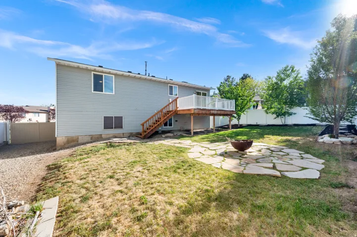 Spacious backyard featuring a stone patio with a fire pit, a wooden deck with white railings, and mature trees