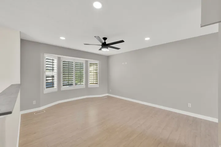 Spacious room featuring light wood-finish flooring, a contemporary black ceiling fan, multiple recessed ceiling lights, and a bay window with white plantation shutters