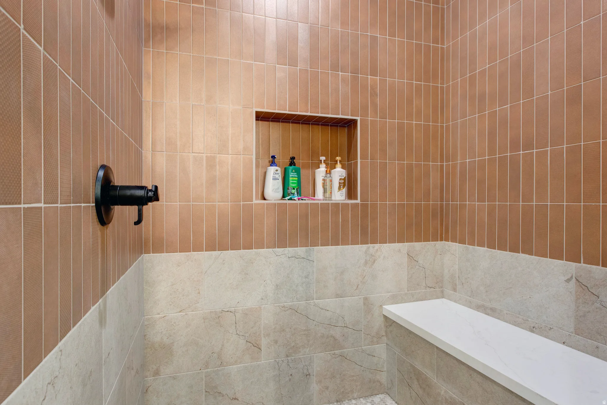 Shower enclosure featuring vertical brown subway tile, lower natural stone-finish wall tiles, an integrated shower niche, a built-in bench with a light-colored surface, and a dark bronze shower fixture
