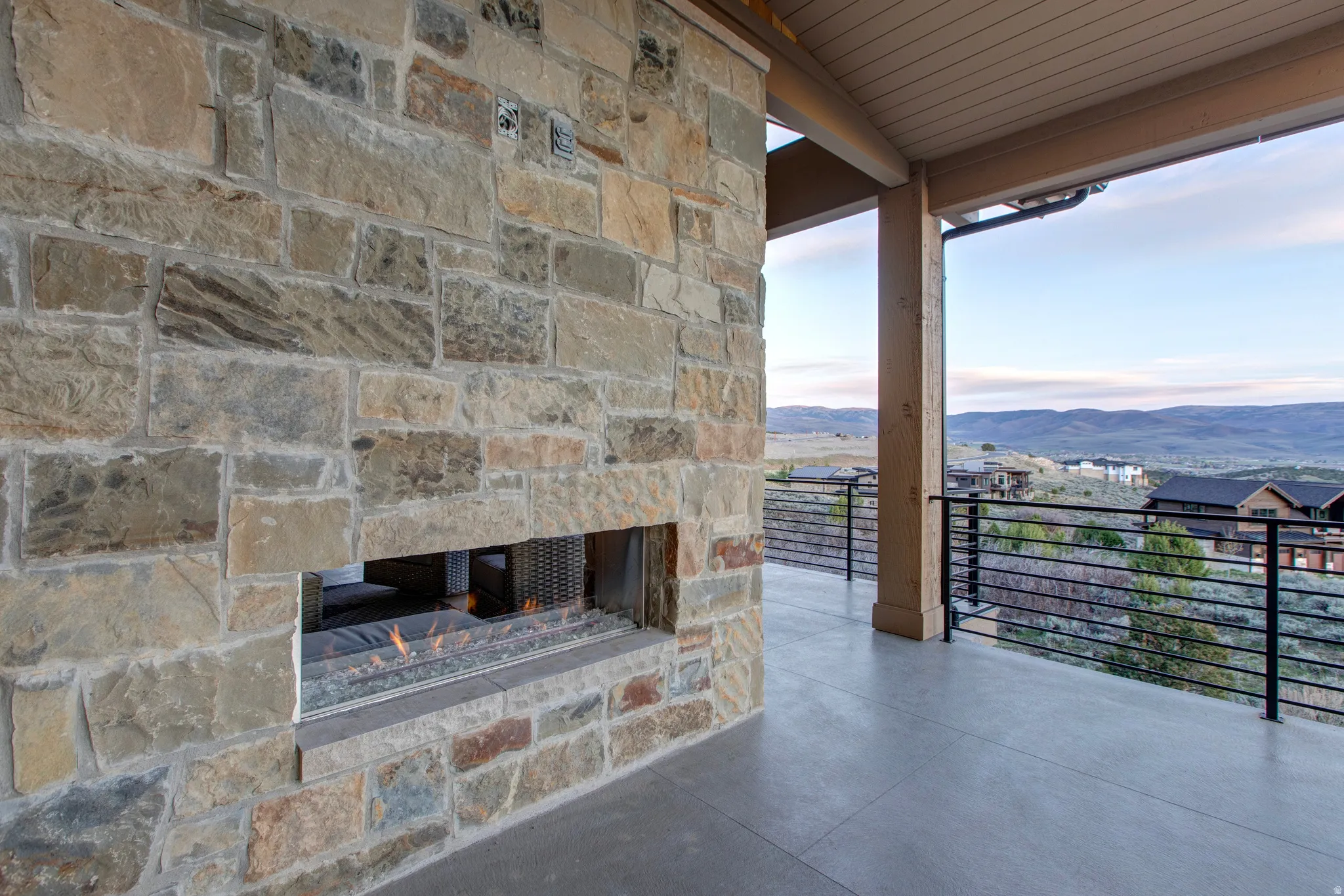 Outdoor patio featuring a stone-clad fireplace with a linear fire feature, concrete flooring, and an open balcony with horizontal metal railings