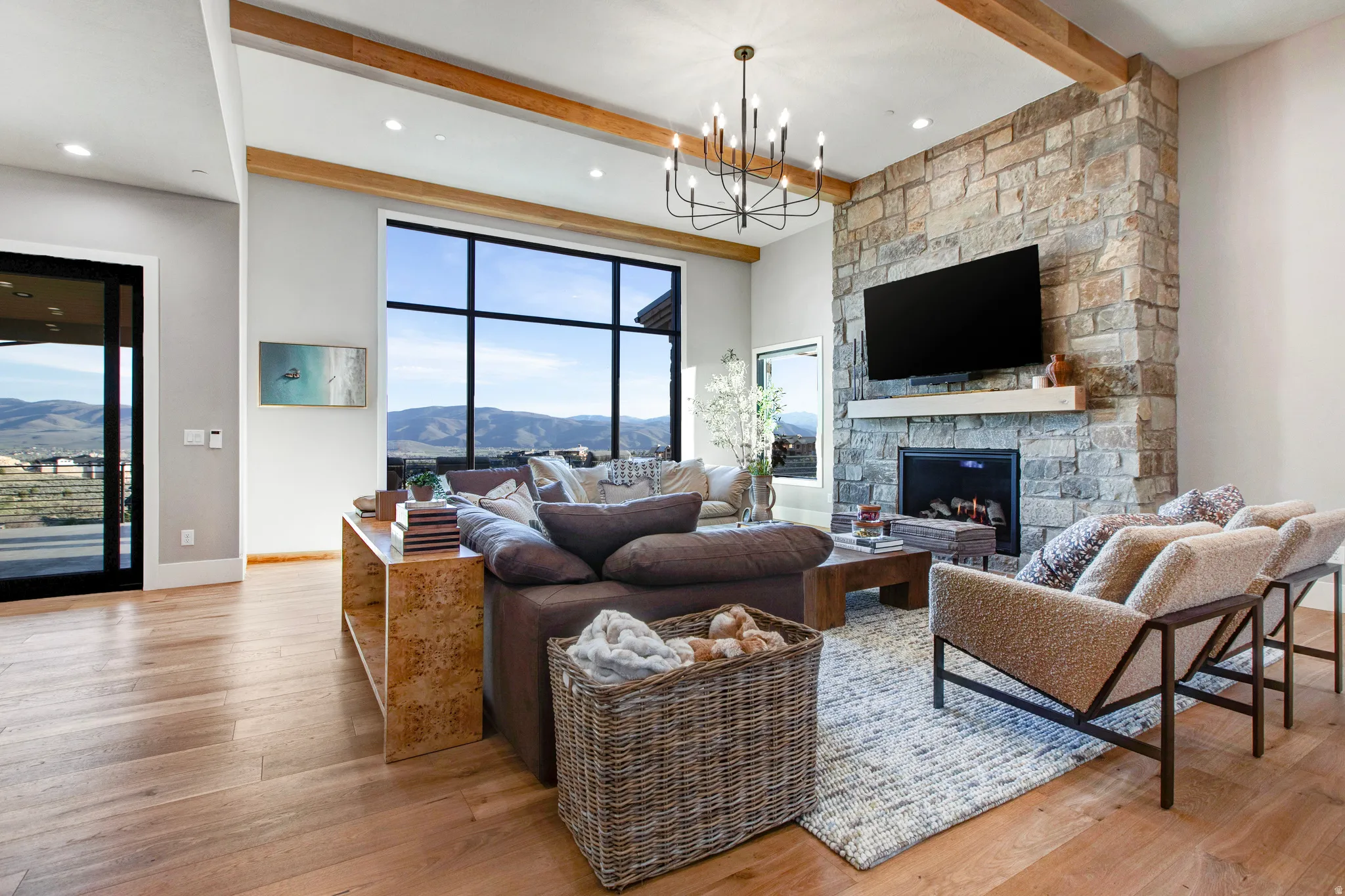 Expansive living area featuring a stone-clad fireplace with mantel, a large picture window showcasing mountainous scenery, wood-finish flooring, exposed ceiling beams, and a modern chandelier