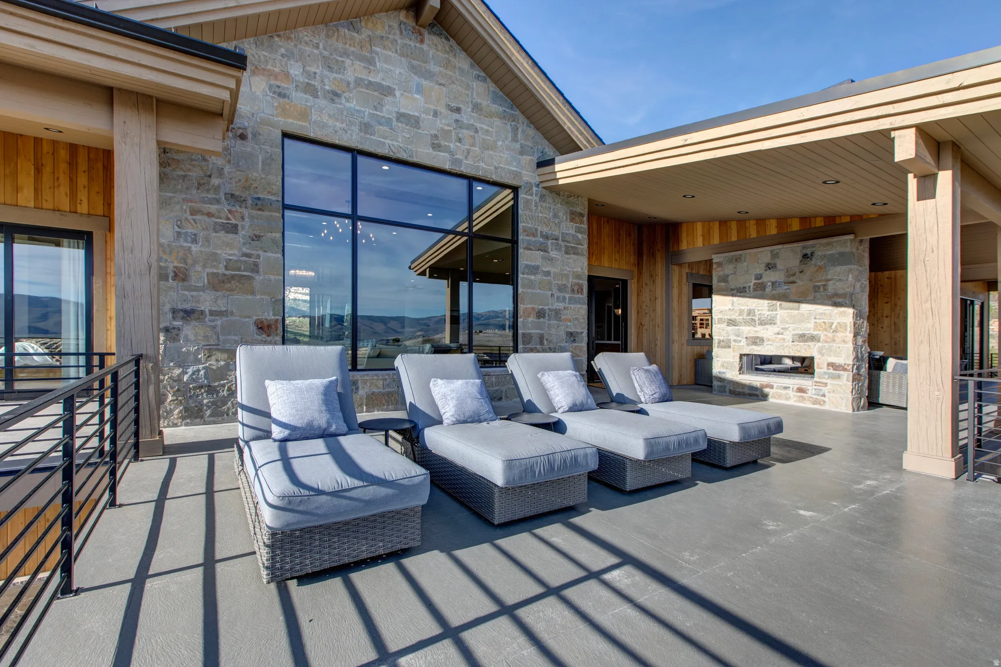 Expansive outdoor patio featuring a stone-clad exterior with large windows, a built-in stone fireplace, a covered section with wood paneling, and a black metal railing