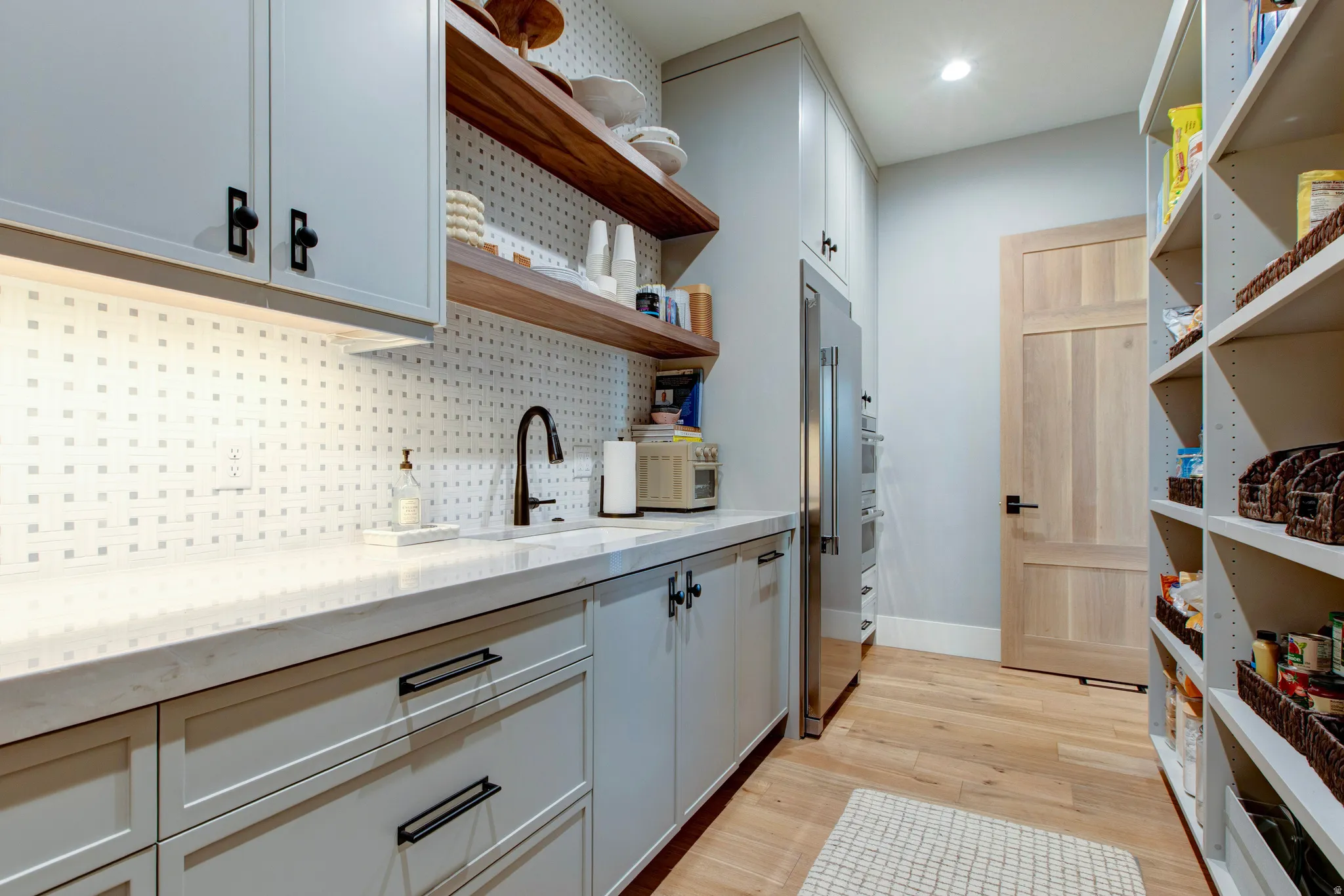 Custom pantry featuring extensive shelving, light wood-finish flooring, a stainless steel refrigerator, and integrated double ovens