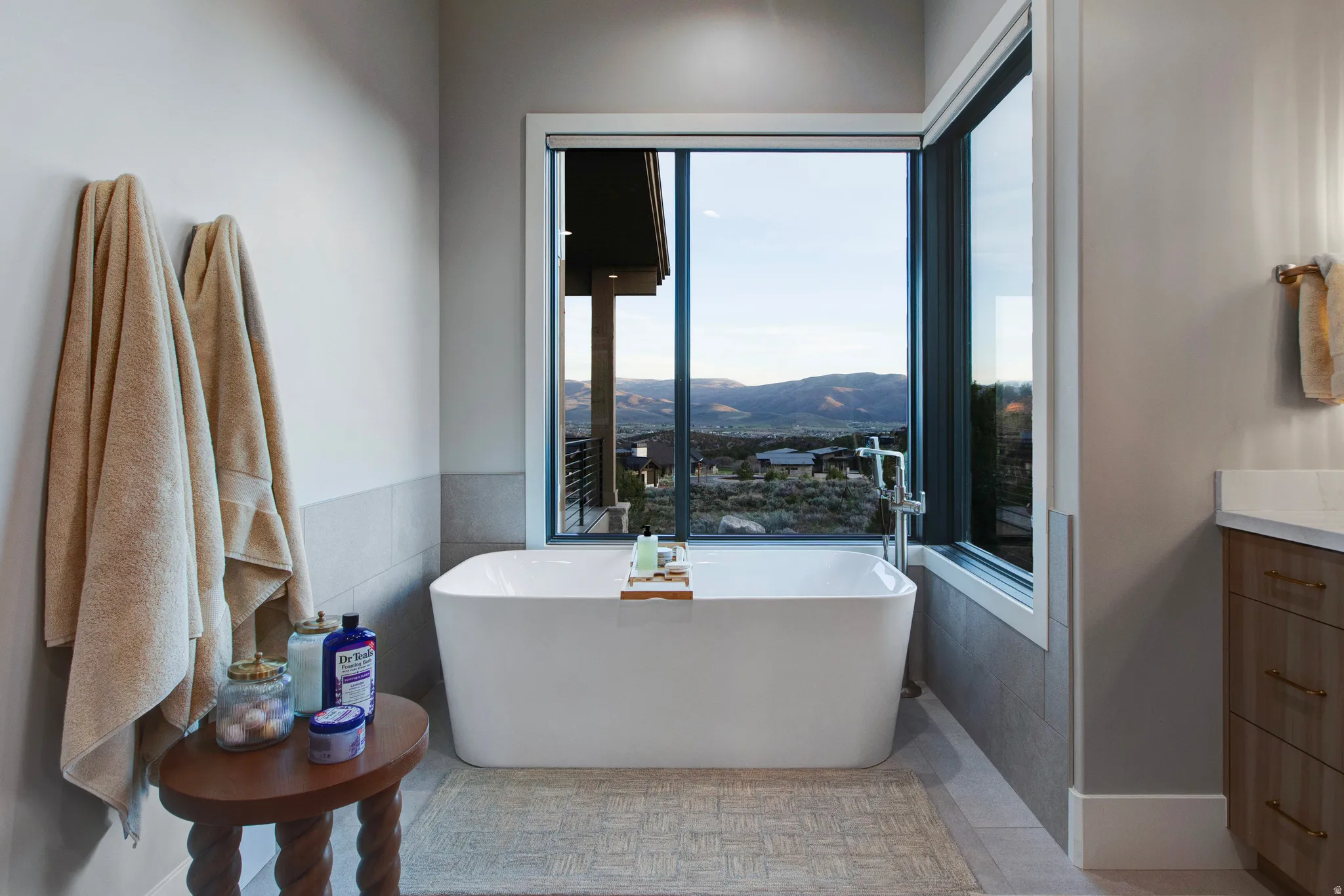 Freestanding soaking tub with floor-mounted faucet, expansive window overlooking mountain scenery, light wood-finish vanity with countertop, and recessed lighting