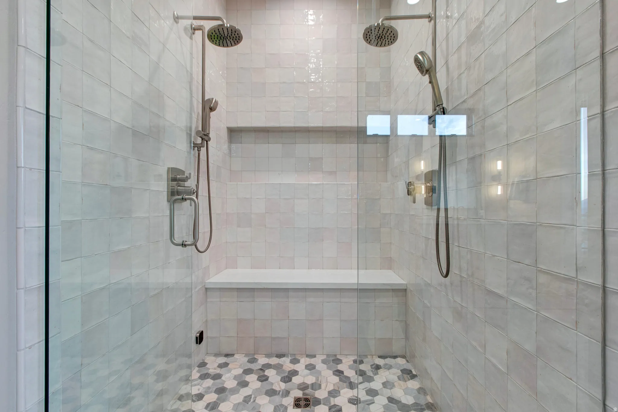 Glass-enclosed shower featuring dual shower heads, a built-in bench, a recessed niche, and hexagonal floor tiling