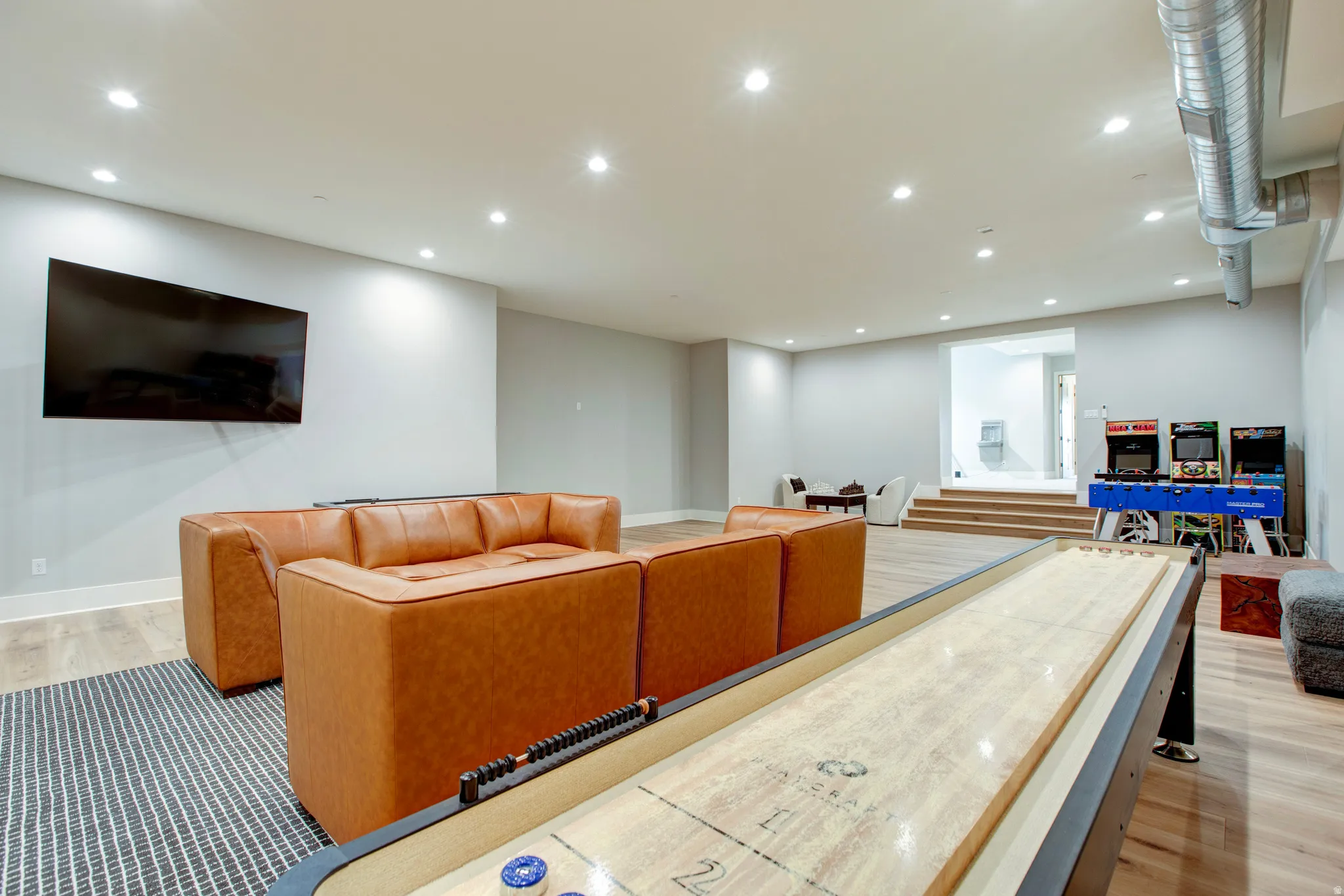 Spacious entertainment area featuring light wood-finish flooring, recessed lighting, and exposed ductwork