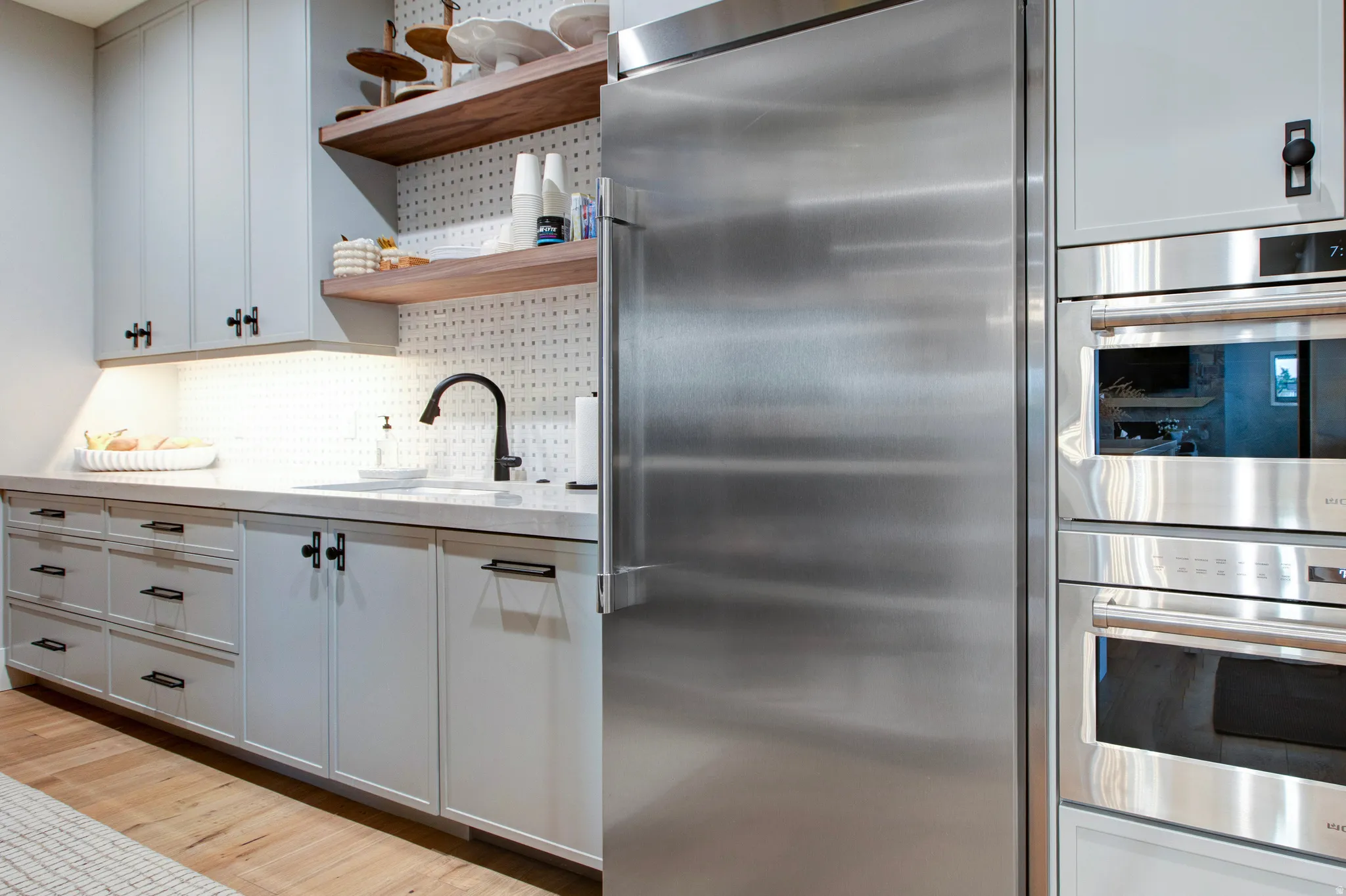Gourmet kitchen featuring a built-in stainless steel refrigerator, double wall ovens, a large sink with a matte black faucet, light grey cabinetry with black hardware, and wood-finish flooring
