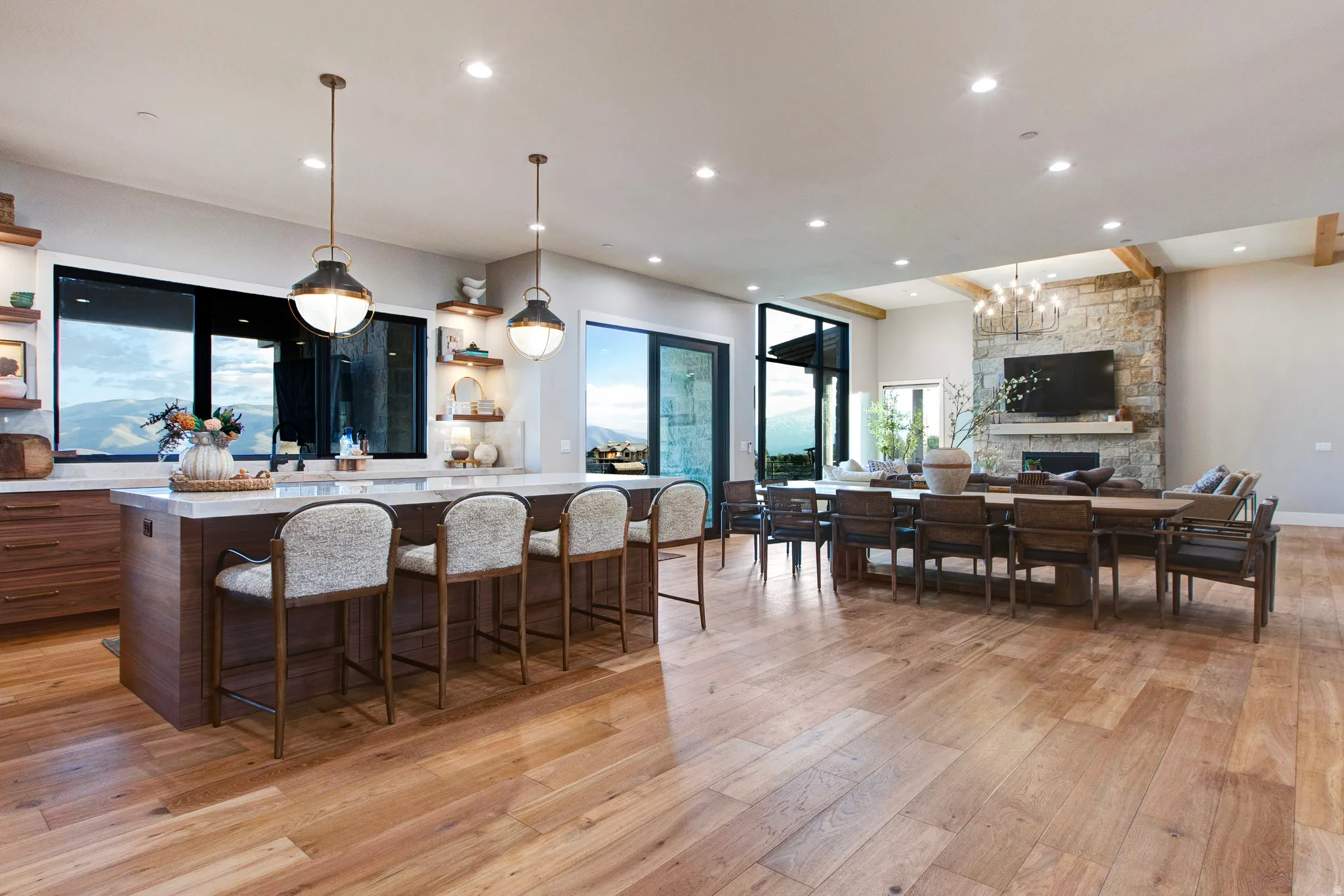 Open-concept living space featuring wood-finish flooring, a kitchen island with pendant lighting, and a stone fireplace with exposed wood beams