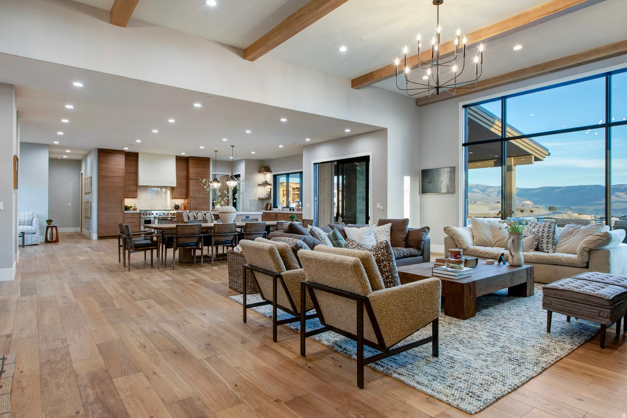 Spacious open-concept living area featuring a vaulted ceiling with exposed wood beams, wood-finish flooring, a contemporary chandelier, a large panoramic window, and recessed lighting throughout