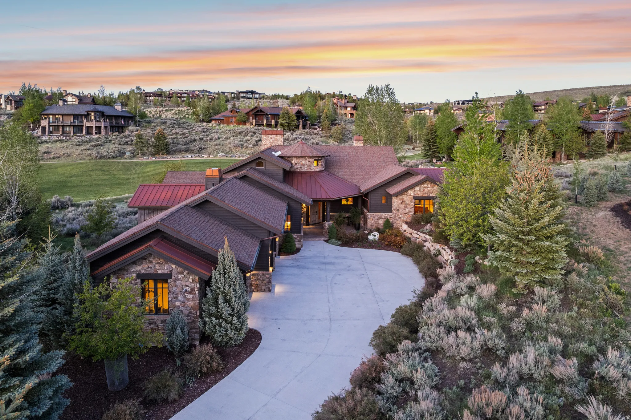 Expansive stone and wood-clad residence featuring a multi-gabled roofline with copper accents, a sweeping paved driveway, and established evergreen landscaping